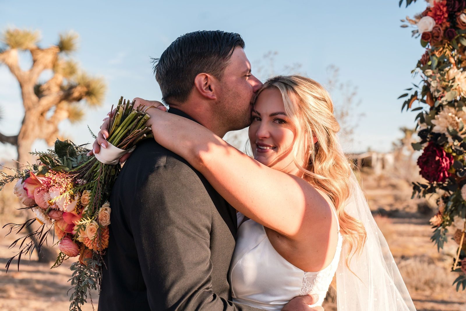 A groom kissing his laughing bride on the cheek, Joshua trees behind them
