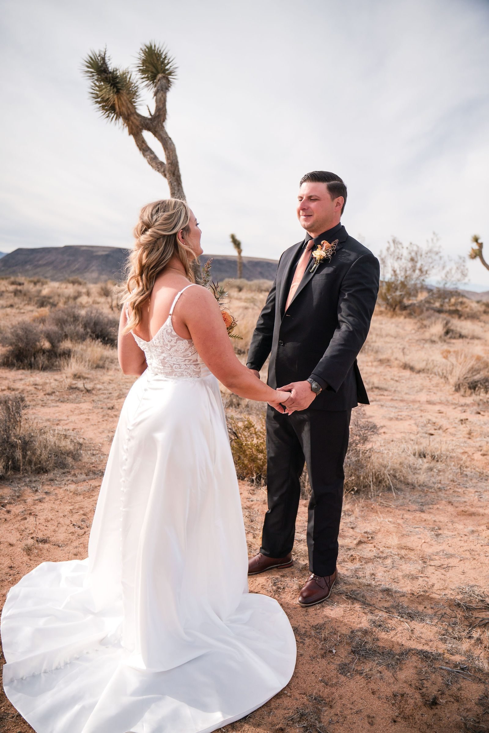 A bride and groom holding hands among Joshua trees under a wide pale sky