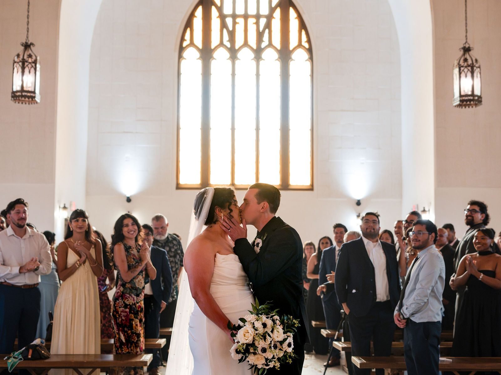 The couple kissing in the aisle as their guests stand on either side under the arched window