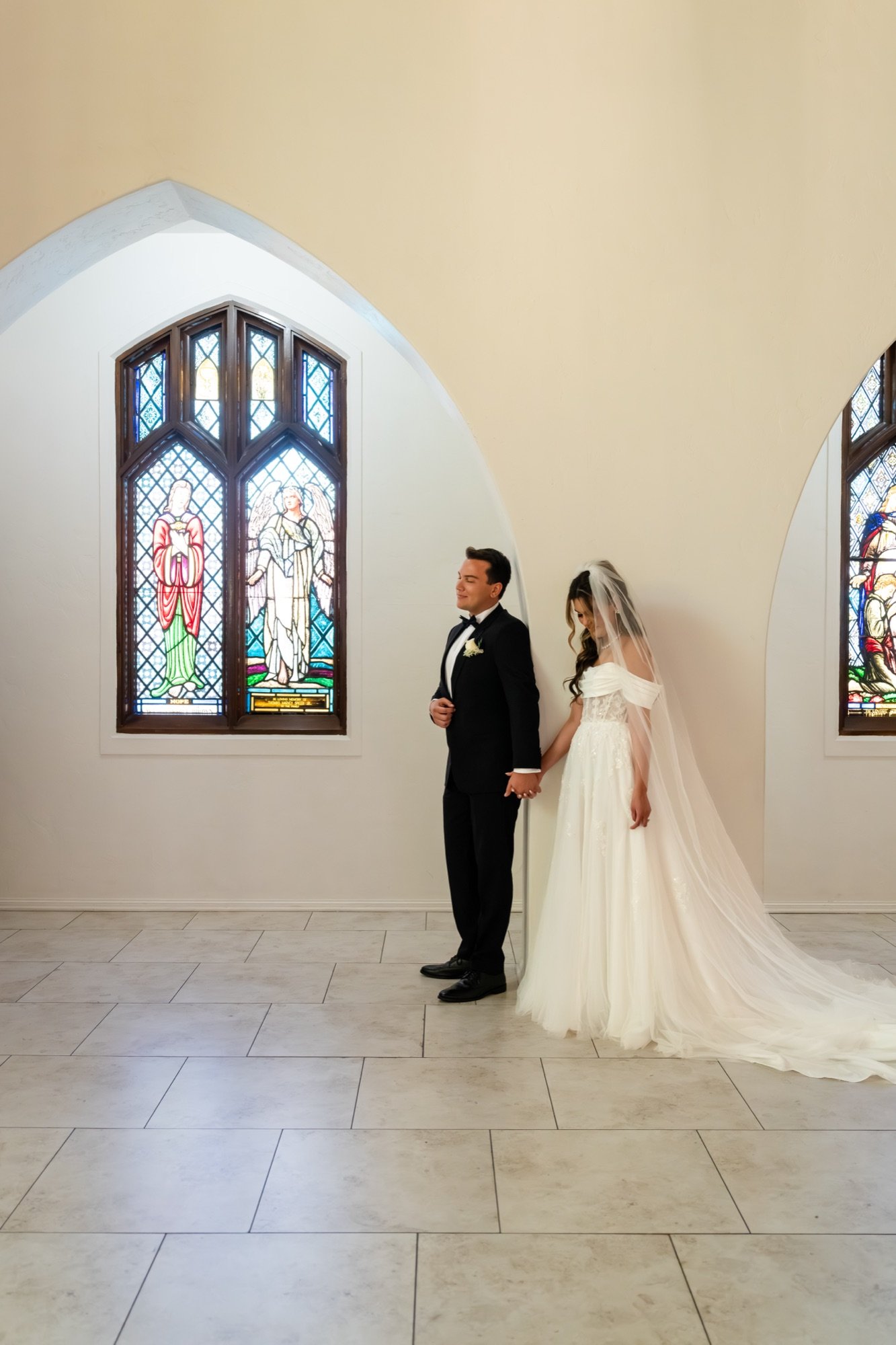 A bride and groom holding hands between two stained glass windows before the ceremony