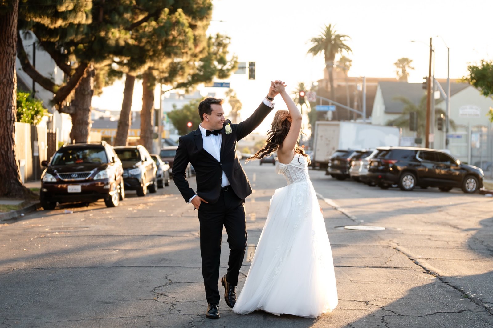 A groom twirling his bride in the street outside at golden hour, palm trees behind them